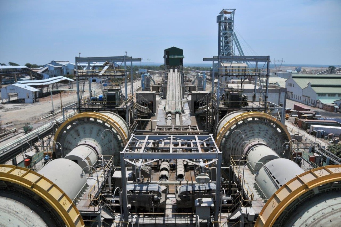 Large-scale mining processing facility featuring massive ball mills or grinding equipment, conveyor systems, and a headframe tower in the background, representing the complete mineral processing chain in modern mining operations.