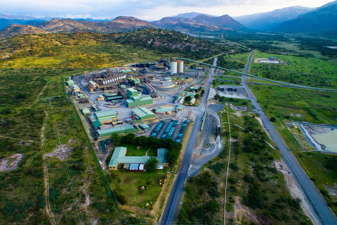 Aerial drone photograph of a comprehensive mining facility showing processing plants with green-roofed buildings, large storage silos, conveyor systems, and road infrastructure set against mountainous terrain.